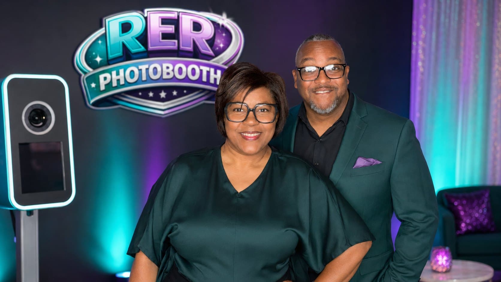 A couple posing beside the digital photo booth station in front of the R.E.R. Photo Booth backdrop